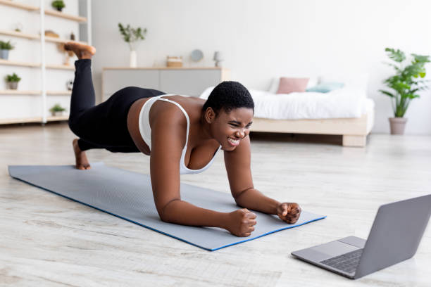 istock Plus size Afro lady working out at home with online personal trainer, using laptop, standing in elbow plank pose 1337895376
