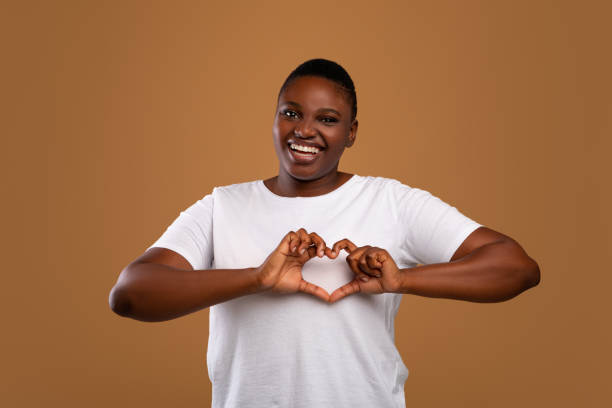 Portrait of casual young black woman shaping hands like heart Kind black chubby lady shaping hands like heart, making love gesture near chest, friendly African American woman expressing kindness and self-love, smiling at camera, standing over brown studio wall huge black woman pictures stock pictures, royalty-free photos & images