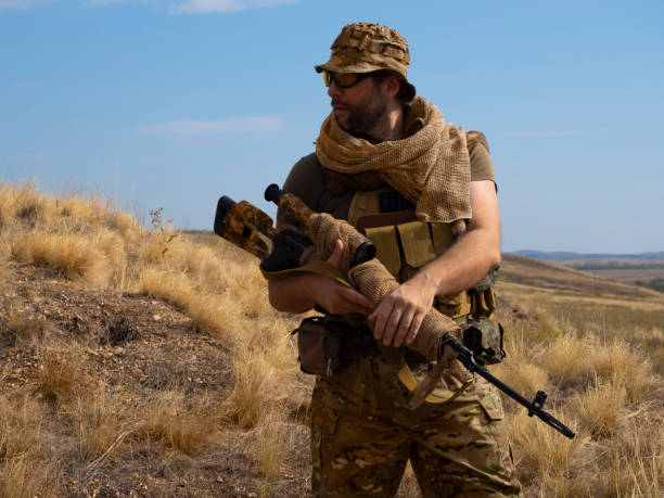 mercenaire tireur d’élite en tenue de camouflage sous le soleil brûlant. il se tient debout avec un fusil et arpente la zone - tireur délite photos et images de collection