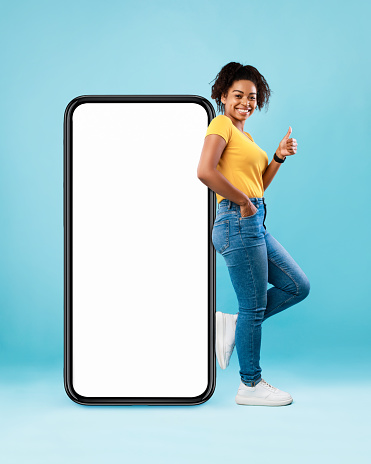 Pretty black lady standing near big smartphone with blank screen, gesturing thumb up on blue background, mockup. Young woman recommending new mobile app, advertising website, free space