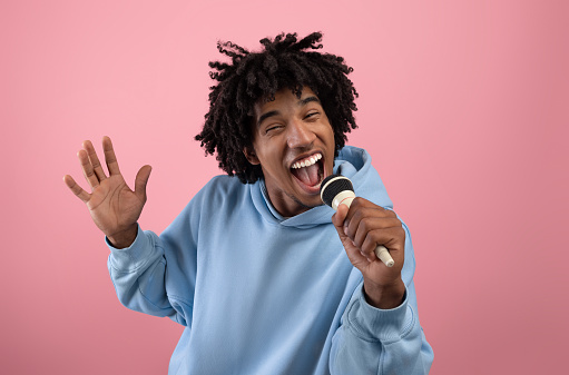 Rock star. Portrait of cool black teen guy singing song, using microphone, performing karaoke on pink studio background. Carefree African American adolescent singer giving live music concert