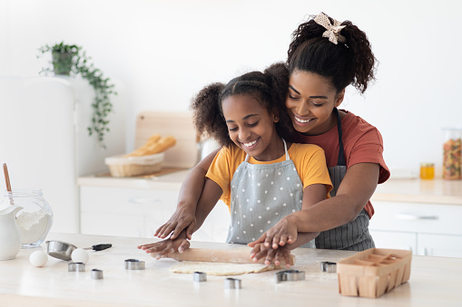 Cheerful african american mother and daughter rolling dough for homemade pastry and smiling, having fun while baking together at home, wearing aprons, kitchen interior, copy space