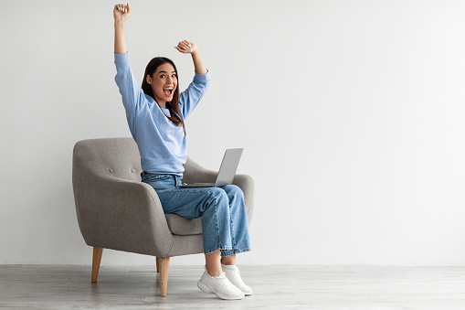 Full length of emotional Caucasian woman with laptop raising hands in excitement, sitting in armchair, receiving great online news, winning lottery, celebrating success or achievement, copy space