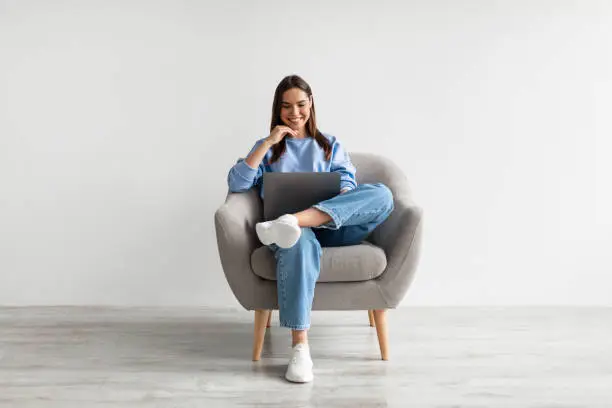 Millennial Caucasian lady in casual wear using laptop, sitting in armchair, having online conference against white studio wall. Young woman communicating online, taking part in webinar
