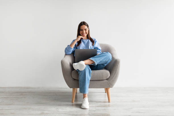 senhora caucasiana millennial em uso casual usando laptop, sentado em poltrona, tendo conferência on-line contra parede branca - laptop isolated computer white - fotografias e filmes do acervo