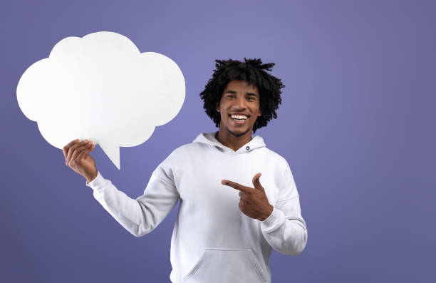istock Funky black teen guy pointing at empty speech bubble, smiling at camera on violet studio background, mockup 1337907295