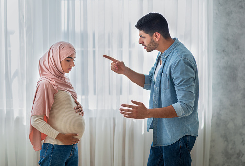 Relationship problems. Aggressive arab man arguing with his upset pregnant muslim wife at home, standing near window, side view. Stressed expectant lady in hijab feeling unwell, touching belly