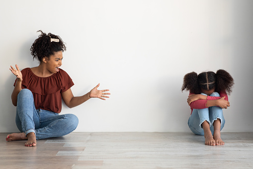 Domestic violence, kids abuse at home concept. Mad black lady abusive mother emotionally gesturing and shouting at her crying female kid, sitting together on floor over empty wall, copy space