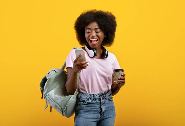 Happy black lady with backpack, headphones, cellphone and takeaway coffee standing over yellow background Online education. Happy black lady with backpack, headphones, cellphone and takeaway coffee standing over yellow studio background. Young female student ready for classes education student mobile phone university stock pictures, royalty-free photos & images