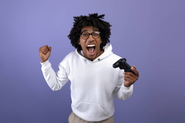 triumfujący czarny nastolatek z joystickiem krzyczący z podniecenia, grający w gry wideo, wykonujący gest tak - gamer zdjęcia i obrazy z banku zdjęć
