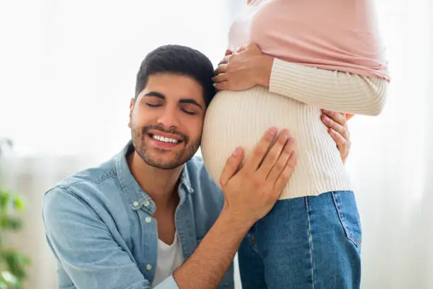 Photo of Delighted eastern man embracing tummy of his pregnant wife, listening baby kicks, tenderly touching belly of his spouse