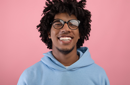 Portrait of joyful black teenager with pleasant smile wearing glasses, looking at camera on pink studio background. Cool African American teen guy with curly hair feeling happy