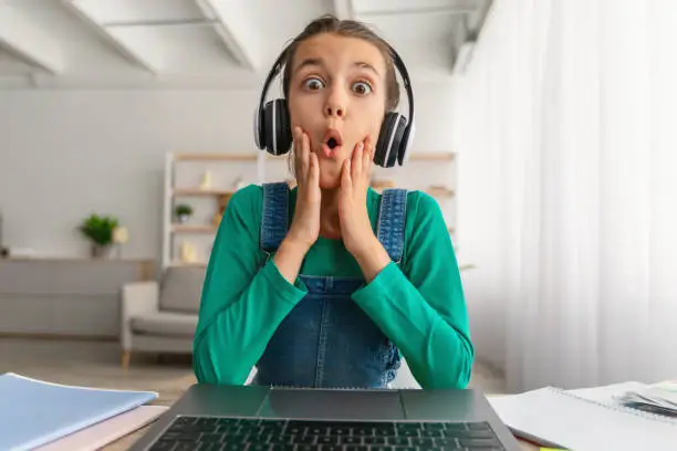 Photo of Surprised girl sitting at desk, having video call wearing headset