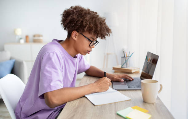 istock Focused black teenager in glasses studying online from home, writing in notebook, using laptop pc at table 1337913232