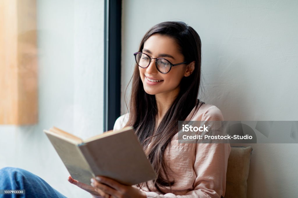 Leisure, education, favorite hobby concept. Cheerful woman reading book in cafe, sitting near window, free space Leisure, education, favorite hobby concept. Cheerful woman reading favorite book in cafe, sitting near window, free space. Happy lady spending time with pleasure Reading Stock Photo