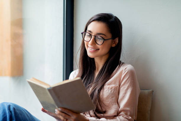 leisure, education, favorite hobby concept. cheerful woman reading book in cafe, sitting near window, free space - woman with glasses reading a book imagens e fotografias de stock