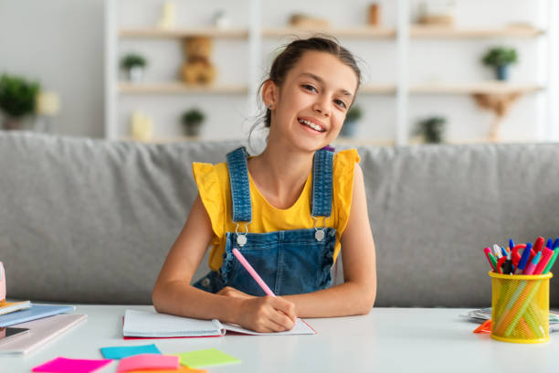 istock Girl sitting at table, writing and posing at camera 1337913470