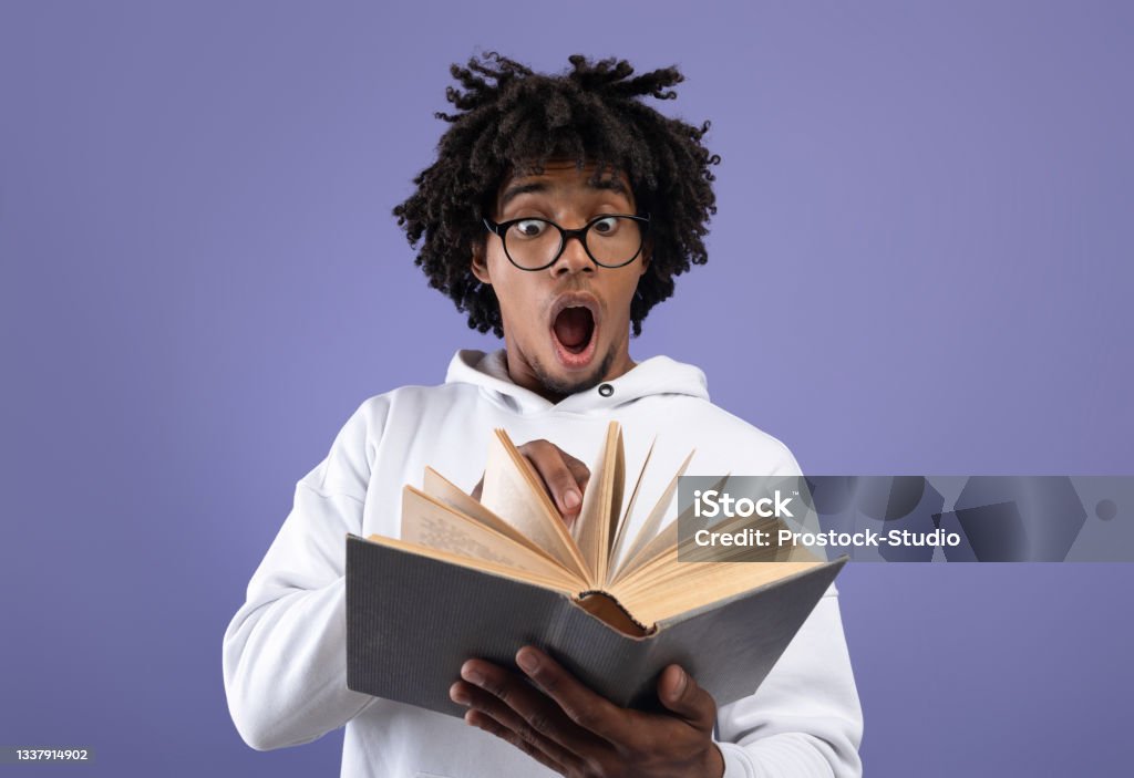 Black teen student looking at textbook in shock, afraid of too much homework on violet studio background Black teen student looking at textbook in shock, afraid of too much homework on violet studio background. Emotional African American teenager studying for difficult college exam, reading book Book Stock Photo
