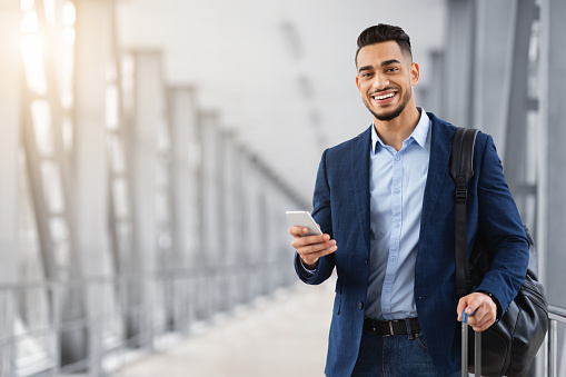 Portrait Of Young Middle Eastern Man With Smartphone In Hand Waiting At Airport, Happy Smiling Arab Businessman Booking Hotel Online Or Browsing Internet On Mobile Phone While Standing In Terminal
