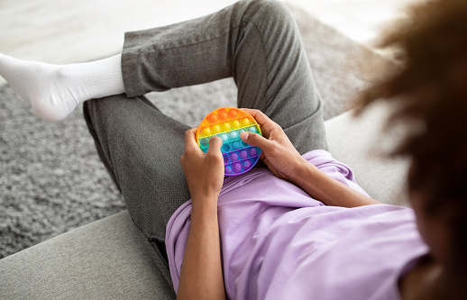 Unrecognizable black teenager playing with POP IT toy, using antistress sensory plaything, sitting on sofa at home