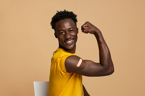 Happy Black Guy Showing Arm With Adhesive Bandage After Covid-19 Vaccination Shot, Vaccinated Young African American Man Demonstrating Biceps With Plaster, Sitting Over Beige Studio Background