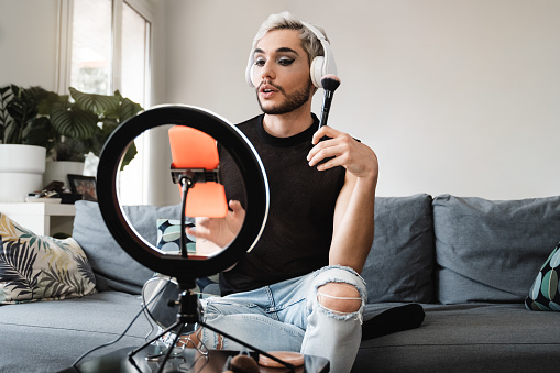 Gay man streaming online make up video tutorial with mobile phone indoors at home - Lgbt, drag queen, technology trendy concept - Focus in face