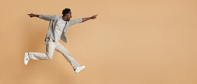 Crazy Sales. Excited black guy jumping up in air and pointing aside at copy space on beige background, emotional young african man demonstrating free place for design or advertisement, panorama