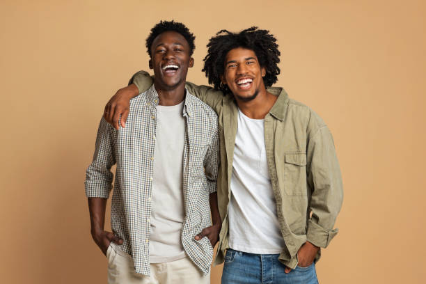 Portrait Of Two Happy Black Guys Embracing While Posing Over Beige Background Portrait Of Two Happy Black Guys Embracing While Posing Over Beige Background, Cheerful Young African American Friends In Stylish Clothes Having Fun In Studio, Laughing At Camera, Copy Space men stock pictures, royalty-free photos & images