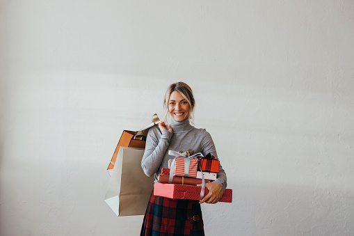 Smiling female holding wrapped gift boxes and paper bags in hands and looking at camera while standing over a white wall