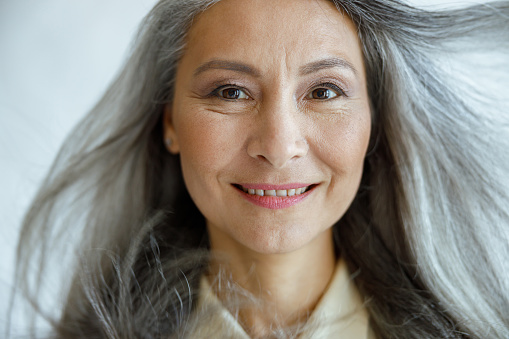 Positive middle aged Asian woman with flying hoary hair looks into camera on light grey background in studio closeup. Mature beauty lifestyle