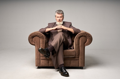 Full length shot of brutal mature businessman with beard wearing elegant suit looking at camera, sitting with hands crossed in armchair while posing isolated over light background. Front view