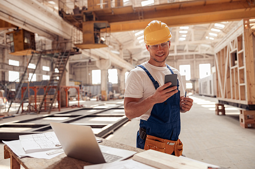 Joyful young man construction worker holding mobile phone and smiling while standing by the table with laptop