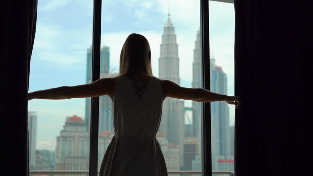 Superslowmotion shot of a silhouette of a successful rich woman opening the curtains of a window overlooking the city center with skyscrapers.