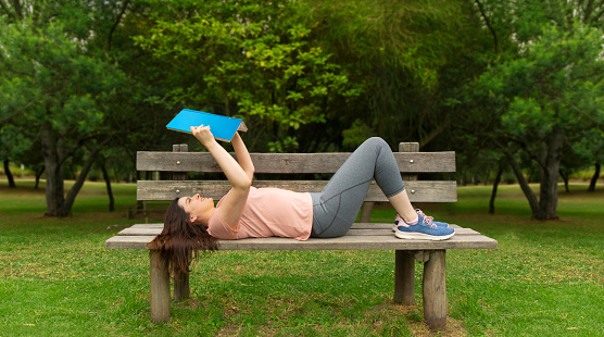 Beautiful Hispanic young woman in sportswear lying on the wooden bench in the middle of a park without people reading a blue book against a background of green trees