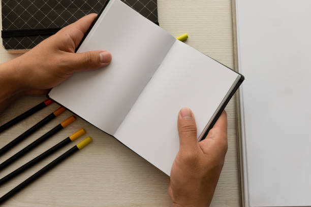 hands holding an open notebook with blank sheets and colored pencils in the background on a desk, office instruments in workspace, detail of objects in studio stock photo