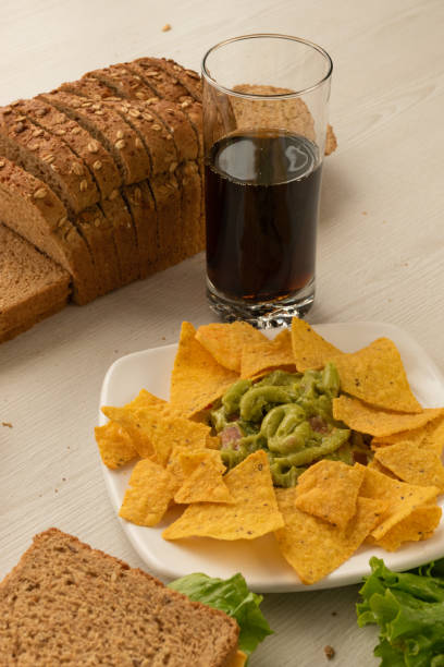 table with a plate of crunchy nachos accompanied by guacamoles, sandwich with cheese, lettuce and tomato, a mold of sliced whole wheat bread and a glass with drink, snack - carbohydrate freshness food and drink studio shot imagens e fotografias de stock