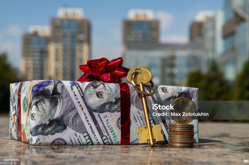 1 American dollar coin, golden key  and a gift wrapped in gift paper depicting 100 American dollars against the background of modern buildings American One Hundred Dollar Bill Stock Photo