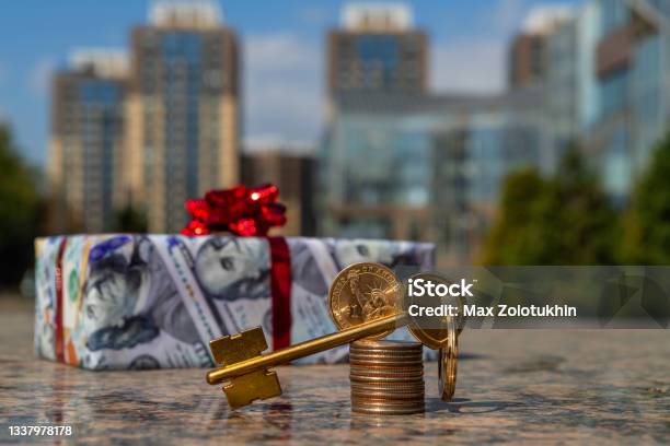 1 American Dollar Coin Golden Key And A Gift Wrapped In Gift Paper Depicting 100 American Dollars Against The Background Of Modern Buildings Stock Photo - Download Image Now