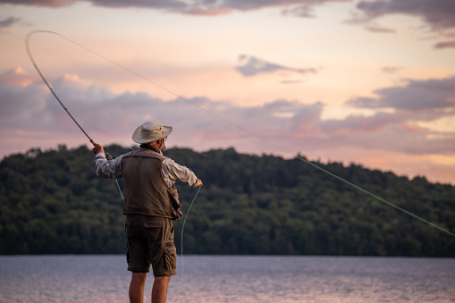 Senior Man Fly-Fishing at Sunset at the lake. He is standing on a pier at Lac St-Joseph, Quebec, Canada.