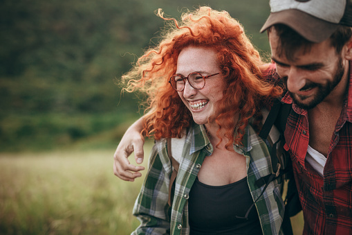 Spontaneous photo of a beautiful young woman, a ginger with a curly hair enjoying an outdoors walks in nature, on a beautiful sunny day, with her best friend, a cool looking bearded guy, hugging her, both radiating happiness with a big smiles. Both being casually dressed, wearing a red and green plaid shirts, enjoying