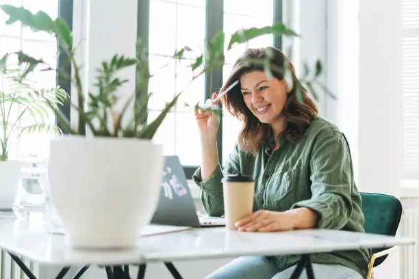 Photo of Young smiling brunette woman plus size working at laptop on table with house plant in the bright modern office