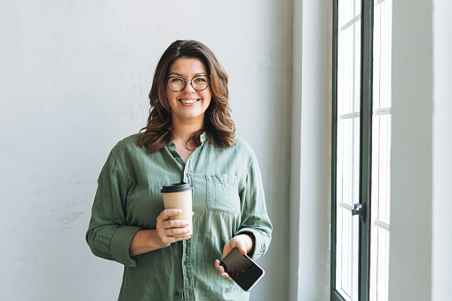 Portrait of Young smiling brunette woman plus size near window with paper cup of coffee and smartphone in hands in the bright modern office
