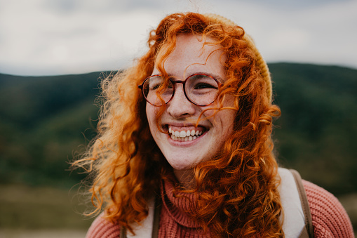 Spontaneous portrait of a beautiful, cheerful, and authentic young adult woman. A curly redhead with a freckles wearing eyeglasses and a peach neck roll sweater with a backpack.She radiates happiness and showing a big toothy smile