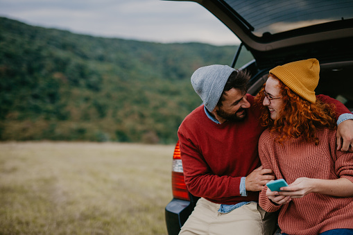 Close up of young couple grabbing their hiking gear from the car and going hiking. Smiling female hiker showing her husband something on a smartphone.