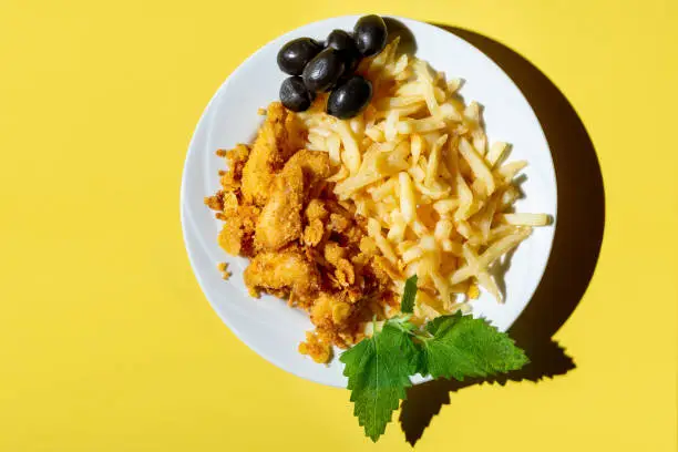 Photo of Nuggets with sauce in a plate with French fries and olives on a yellow background with red napkins. hard shadows.