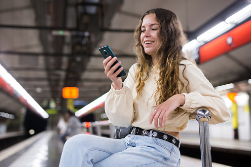 Portrait of a positive girl sitting on a platform bench in the subway, texting with friends on a mobile phone while \nwaiting for the train