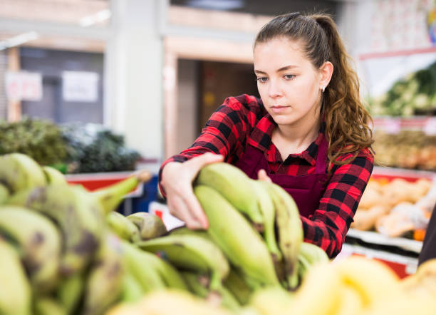 istock Young female shopping assistant in apron selling bananas 1338007764