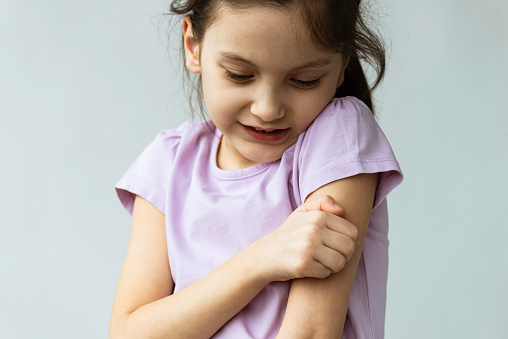Little girl is scratching her arm in front of gray background.