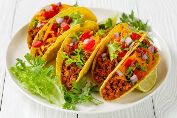 istock Ground Beef Tacos with shredded cheddar cheese, fresh lettuce, tomato, onion on a white plate with lime wedges, horizontal view from above, mexican cuisine, close-up 1338013654