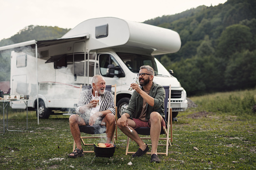 A mature man with senior father talking at campsite outdoors, barbecue on caravan holiday trip.